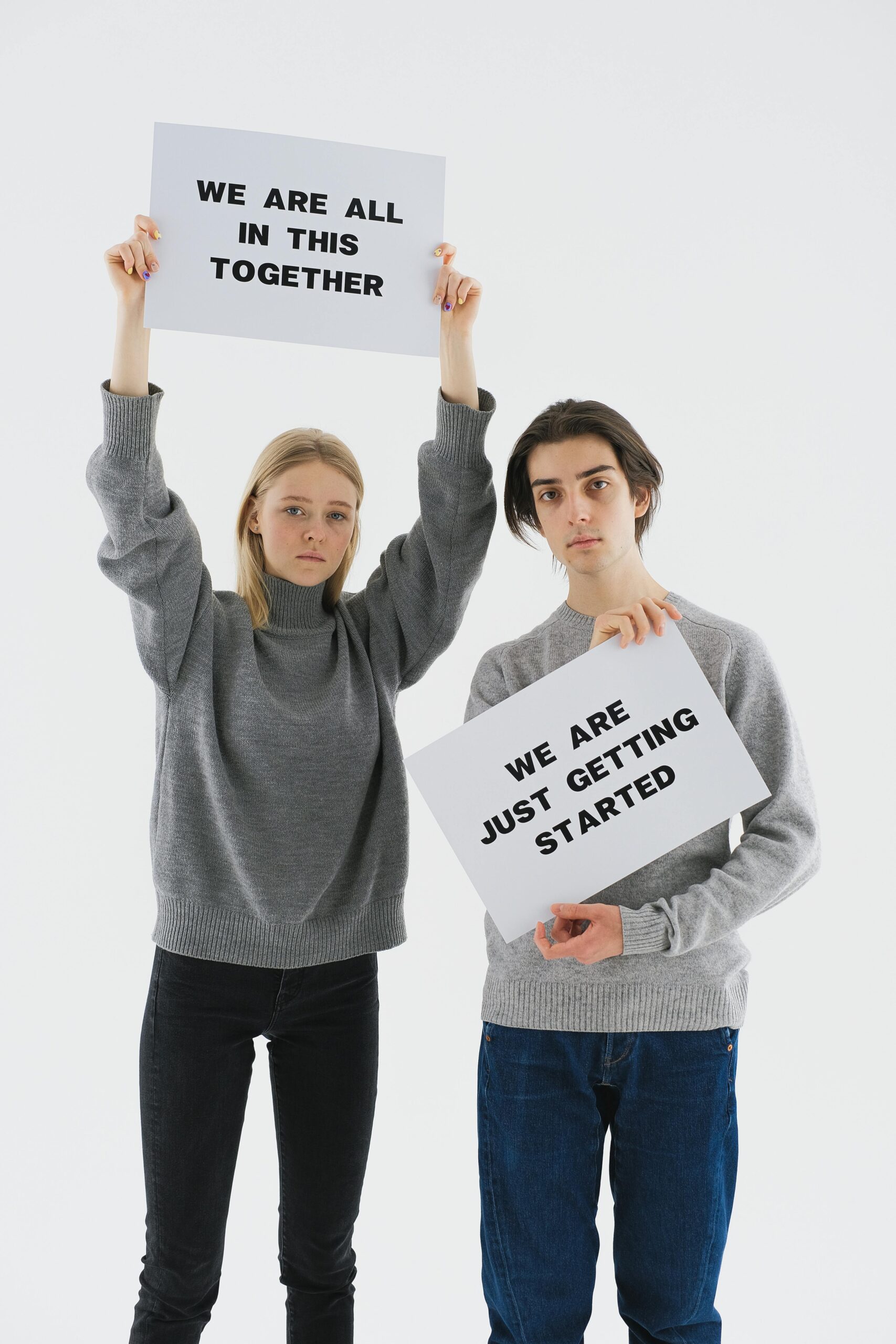 Two young adults in gray sweaters holding motivational signs against a white background.